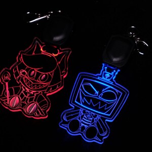 May include: Two illuminated keychains with cartoon character designs. One keychain glows red, featuring a character with sharp teeth and a staff. The other glows blue, depicting a robot-like character with a menacing grin. Both have black key rings.