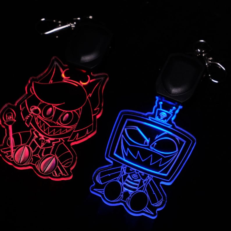 May include: Two illuminated keychains with cartoon character designs. One keychain glows red, featuring a character with sharp teeth and a staff. The other glows blue, depicting a robot-like character with a menacing grin. Both have black key rings.