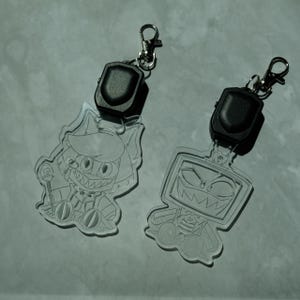 May include: Two clear acrylic keychains with cartoon character designs. Each keychain has a black, rectangular light and a silver clasp. One features a cat-like character, the other a TV-headed figure, both with sharp teeth.