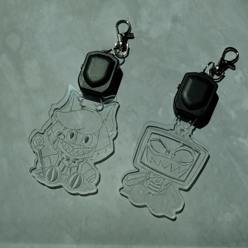 May include: Two clear acrylic keychains with cartoon character designs. Each keychain has a black, rectangular light and a silver clasp. One features a cat-like character, the other a TV-headed figure, both with sharp teeth.