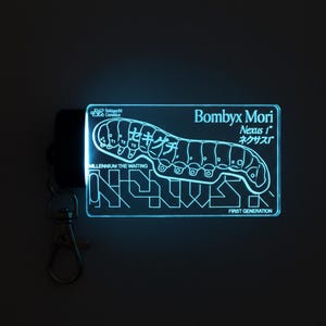 May include: A rectangular, light-up acrylic keychain featuring a glowing blue outline of a silkworm. The text reads Bombyx Mori, Nexus 1, Millennium The Waiting, and First Generation, along with Japanese characters and Sekiguchi Genetics branding.