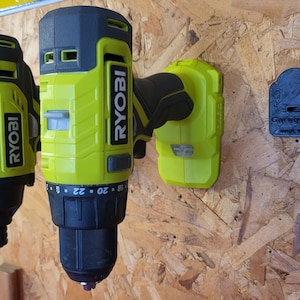 May include: Two yellow and black Ryobi power drills mounted on a wooden surface. The drills are in a vertical position. A black 3D-printed holder with the text "GarageGuy 3D" is also mounted on the surface.