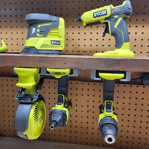 May include: A collection of Ryobi power tools in yellow and gray, including a sander, glue gun, impact driver, drill, and a fan. The tools are displayed on a wooden shelf and hanging from a rack against a pegboard.