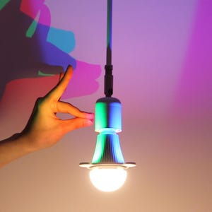 May include: A contemporary LED light bulb with a white base and a colourful, gradient-lit top. The light is suspended from a dark grey cord. The image features a hand interacting with the light, and colourful shadows on the wall.