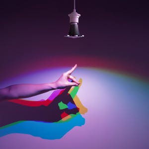 May include: A hand gestures towards a colorful shadow, with a light bulb fixture hanging from above. The shadow is a combination of red, green, blue, and yellow. The background is a gradient of purple and pink.