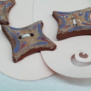 May include: Three ceramic buttons shaped like a four-pointed star. The buttons are brown with a beige glaze and have a blue and pink design. The buttons have two holes for sewing.