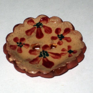 May include: A round, ceramic button with a scalloped edge. The button is decorated with a floral pattern in shades of red and brown on a beige background.