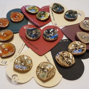 Puede incluir: Surtido de botones de cerámica hechos a mano en varios colores y patrones, incluyendo diseños florales, moteados y abstractos. Los botones se muestran en tarjetas con forma de corazón con la dirección del sitio web "www.mimsical.co.uk" impresa en ellas.