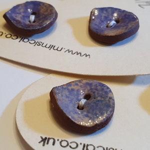 May include: Three ceramic buttons in a teardrop shape with a purple and brown glaze. The buttons have a white thread hole in the center.