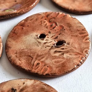 May include: Two-hole brown ceramic button with a white and orange marbled design. The button has a raised, textured pattern.