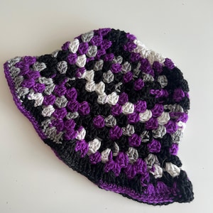 May include: A handmade crochet hat in a granny square pattern. The hat features a color scheme of black, purple, white, and gray. The hat is a unique, colorful accessory.
