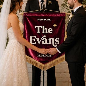 May include: A deep burgundy velvet banner with gold fringe trim, featuring the white embroidered text NEWYORK, The Evans, and the date 25.06.2026, accented by a small gold star graphic, held between two people during a wedding ceremony.