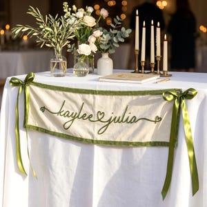 Op de afbeelding: Een crèmekleurige stoffen banner met de geborduurde groene tekst "kaylee julia" en hartjes, afgezet met een groene fluwelen rand en aan beide zijden bevestigd met groene satijnen linten, getoond op een wit tafelkleed.