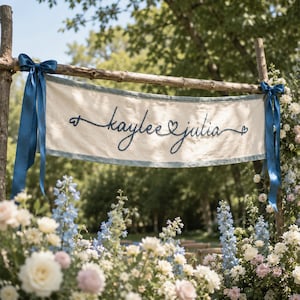 May include: A rectangular off-white fabric banner featuring the embroidered dark blue text "kaylee julia" with heart accents. The banner is suspended from a rustic wooden frame by dark blue satin ribbons tied at each end of the top bar.