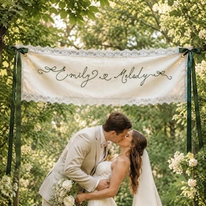 Könnte beinhalten: Ein rechteckiges Banner aus cremefarbenem Stoff mit dem in schwarzer Schreibschrift gestickten Text Emily & Melody. Das Banner ist an den oberen und unteren Kanten mit weißer Spitze verziert und wird von dunkelgrünen Satinbändern gehalten.