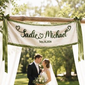 以下が含まれることがあります： オフホワイトの布製長方形バナー。ダークグリーンの刺繍で「Sadie & Michael」と日付「Sep, 26, 2026」が記されています。バナーにはダークグリーンの縁取りが施され、ダークグリーンのサテンリボンで木の梁に結び付けられています。