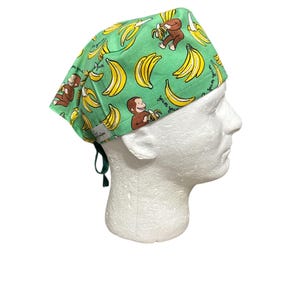 May include: A green surgical scrub cap featuring a pattern of yellow bananas and brown monkeys. The cap has a tie in the back and the words "yum yum" are printed on the fabric. The cap is displayed on a white mannequin head.