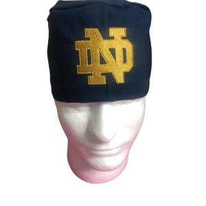 May include: A navy blue beanie-style hat with a gold embroidered "ND" logo. The hat is displayed on a white mannequin head. The hat is a sports-themed accessory.