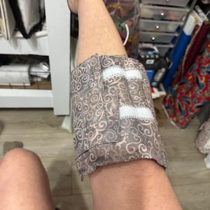 May include: A patterned fabric knee wrap in shades of gray and tan, secured with white hook-and-loop fasteners. The wrap features a swirling design and is wrapped around a leg.