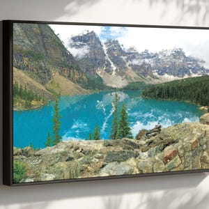 May include: Framed landscape art print featuring a vibrant turquoise lake surrounded by mountains and evergreen trees. The artwork captures a scenic view with snow-capped peaks and a rocky foreground, all within a black frame.