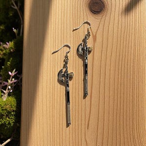 May include: A pair of silver-toned dangle earrings. Each earring features a small, crescent-shaped charm, a decorative cross, and a long, slender, black and silver-toned rod. The earrings are displayed on a wooden surface.