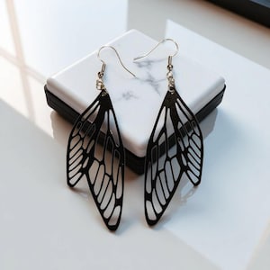 May include: A pair of black, open-work wing-shaped earrings. The earrings have a delicate, filigree design with a hook closure. The earrings are displayed on a white and gray marble surface.