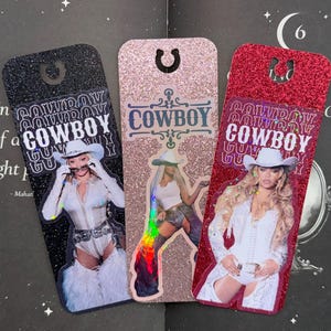 May include: Three glittery bookmarks with images of women in cowboy outfits. The bookmarks are black, pink, and red, with the word "COWBOY" in a stylized font. Each bookmark has a hole at the top for use in a book.