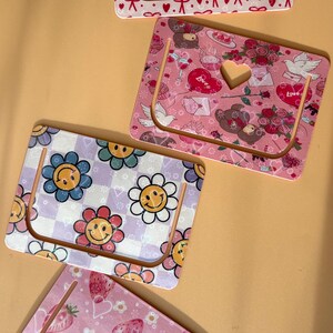 Puede incluir: Juego de cuatro marcapáginas decorativos con varios diseños. Uno presenta lazos rojos sobre fondo blanco, otro tiene un fondo rosa con corazones y ositos de peluche, un tercero tiene flores sonrientes y el último fresas.