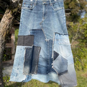 Pode incluir: Uma saia jeans patchwork em vários tons de azul, pendurada em um cabide de madeira. A saia apresenta uma mistura de peças de jeans, incluindo um painel preto e uma seção listrada, com bordas cruas.