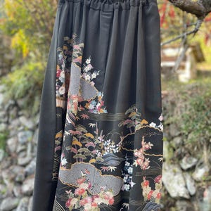 May include: Black skirt with an ornate floral and landscape design. The skirt features a detailed pattern of flowers, trees, and mountains in shades of pink, gold, and green. The skirt is hanging from a wooden rod.