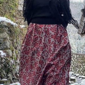 May include: A red and gray floral print skirt with a gathered, voluminous shape. The skirt is paired with a black top, boots, and a black lace shawl. The outfit is worn outdoors in a snowy environment.