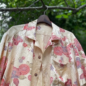May include: A short-sleeved button-down shirt in a light cream color featuring a repeating floral and butterfly pattern in shades of red, purple, and gold. The shirt has a classic collar, a single chest pocket, and dark brown buttons down the front.