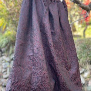 May include: A burgundy skirt with a black, wave-like pattern. The skirt is hanging from a wooden hanger. The fabric has a sheen, and the skirt has a gathered waist and a full shape.