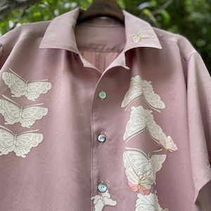 May include: A dusty rose button-down shirt featuring a subtle geometric textured pattern and white butterfly embroidery. The shirt has a classic collar, iridescent buttons, and multiple butterfly designs arranged along the front placket.