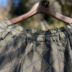 May include: Close-up of a black and gold patterned skirt hanging on a wooden hanger. The fabric has a diamond-shaped design. The skirt is gathered at the waist. The background is blurred, with a tree branch and sunlight visible.