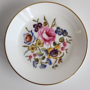 May include: A small, white, round dish with a gold rim. The dish features a floral design with pink, purple, blue, and yellow flowers. The flowers are arranged in a bouquet in the center of the dish.