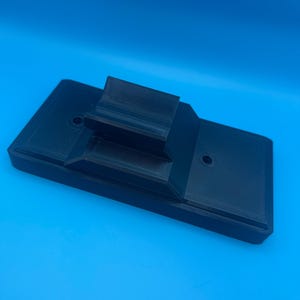 May include: A black, 3D-printed object with a rectangular base and a raised, angled section. Two small holes are visible on the base. The object is set against a bright blue background.