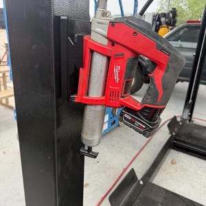 May include: A red and black Milwaukee cordless grease gun attached to a black metal support. The grease gun has a silver cylinder and a black handle with a battery pack attached. The image is taken outdoors.