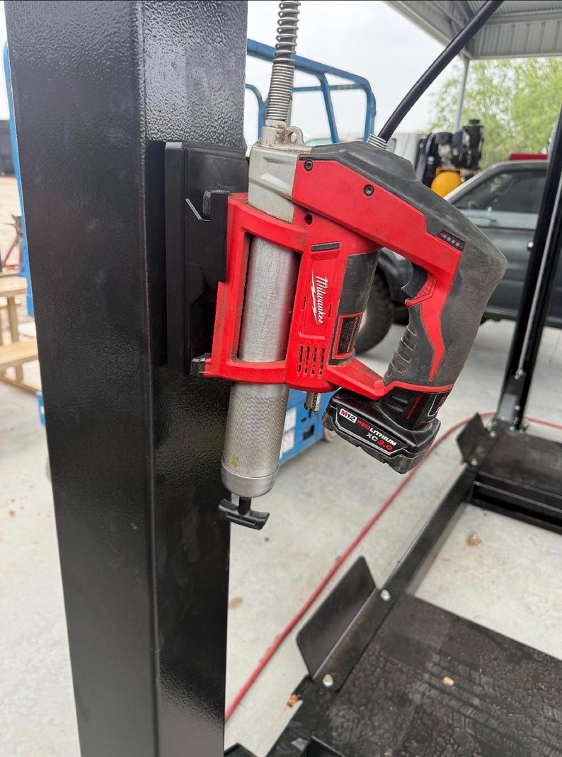 May include: A red and black Milwaukee cordless grease gun attached to a black metal support. The grease gun has a silver cylinder and a black handle with a battery pack attached. The image is taken outdoors.