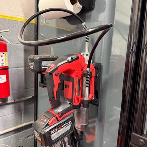 May include: A red and black cordless grease gun with a black hose and a battery pack. A roll of paper towels is mounted above the grease gun. The tools are mounted on a gray metal surface.