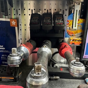 Könnte beinhalten: Eine Sammlung von Werkzeugen für Kraftfahrzeuge, darunter drei Ratschen mit roten und silbernen Komponenten und drei Batterieladegeräte. Die Werkzeuge sind an einer Metalllochwand befestigt, mit einem Hot Wheels-Spielzeugauto im Hintergrund.