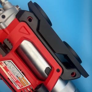 May include: A red and black grease gun with a silver cylinder and a black attachment. The tool has a label with text and is set against a blue background. The grease gun is a power tool.