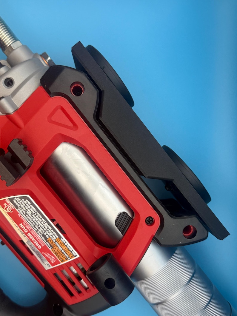 May include: A red and black grease gun with a silver cylinder and a black attachment. The tool has a label with text and is set against a blue background. The grease gun is a power tool.