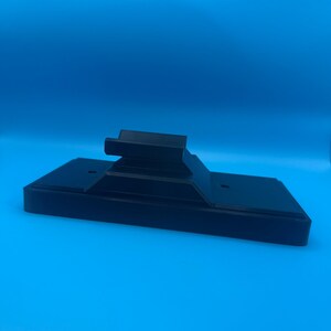 May include: A black, rectangular stand with a raised platform and two small holes. The stand is made of a solid material and is set against a bright blue background. The stand is designed to hold an electronic device.