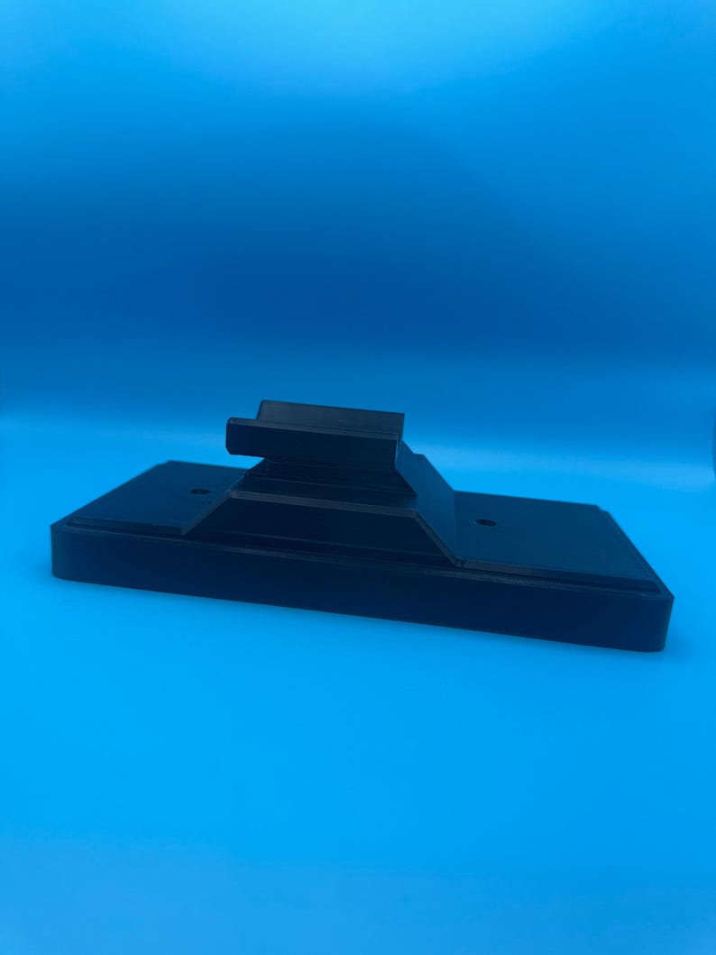 May include: A black, rectangular stand with a raised platform and two small holes. The stand is made of a solid material and is set against a bright blue background. The stand is designed to hold an electronic device.
