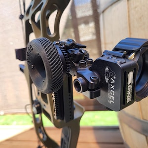 Peut inclure: Gros plan d'une mire de tir à l'arc noire fixée à un arc à poulies vert. La mire comprend une molette de réglage circulaire, un viseur et les logos "AXCEL" et "PINS". L'arc est placé contre un tonneau en bois.