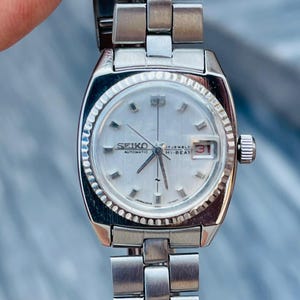 May include: A silver-toned Seiko automatic wristwatch with a rectangular face and a silver band. The watch face is light-colored with the words "SEIKO AUTOMATIC HI-BEAT" and a date display. The watch is approximately 1 inch wide.