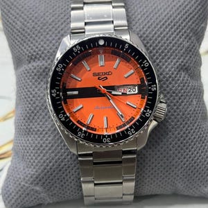 Puede incluir: Un reloj automático Seiko 5 de acero inoxidable con esfera naranja y bisel negro. El reloj tiene una pantalla de día y fecha, y una correa de metal plateado. El reloj se muestra sobre un cojín gris.