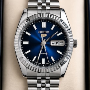 May include: A silver-tone Seiko 5 automatic wristwatch featuring a deep blue sunray dial with luminous baton indices, a fluted bezel, and a stainless steel jubilee-style link bracelet. The dial displays the Seiko logo, day, and date window.