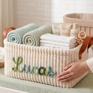 Puede incluir: Una cesta de almacenamiento de pana color crema con el nombre "Lucas" en hilo verde y azul. Contiene mantas enrolladas, paños blancos doblados, productos para el cuidado de la piel del bebé y un juguete de madera. La cesta tiene asas de cuero marrón.